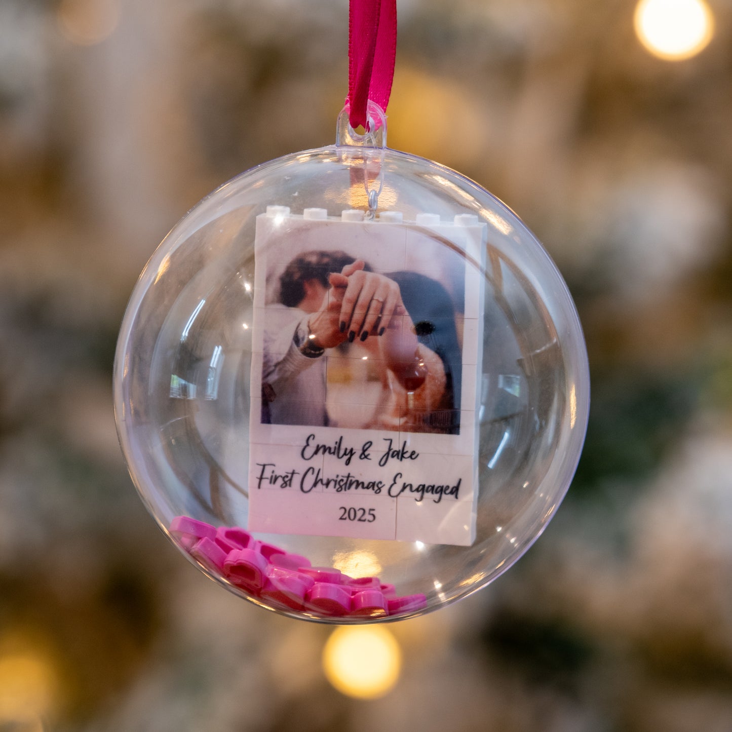 Clear Christmas ornament with a photo of a couple and text, hanging against a blurred festive background.