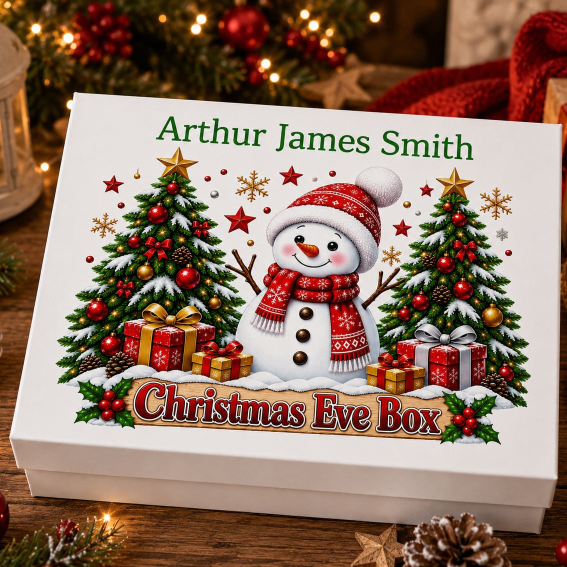 A white rectangular gift box with a Christmas Eve themed illustration on the lid, featuring a snowman in a red hat and scarf between two decorated Christmas trees, with the text 'Arthur James Smith' and 'Christmas Eve Box.' The box is styled on a wooden surface with holiday lights, pinecones, red ornaments, and knitted fabric in the background.