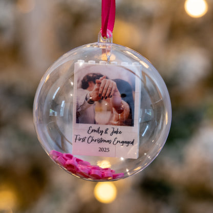 Clear Christmas ornament with a photo of a couple and text, hanging against a blurred festive background.