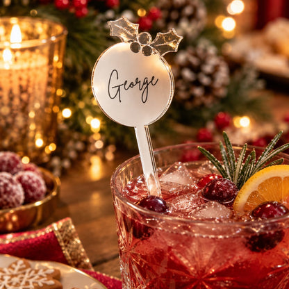 A festive holiday drink in a clear cut-glass tumbler with a personalized place card reading "George" on a metal stirrer, garnished with cranberries, rosemary, and an orange slice, set on a wooden table with candles, pinecones, and cookies in the background.
