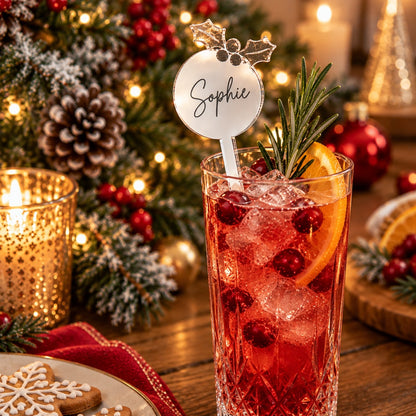 A festive holiday cocktail in a clear cut-glass tumbler with a personalized silver name sign reading 'Sophie' on a pick, garnished with a sprig of rosemary, a cranberry, and an orange slice. The drink sits on a wooden table surrounded by holiday decorations including a lit gold candle holder, pinecones, red ornaments, and gingerbread cookies.