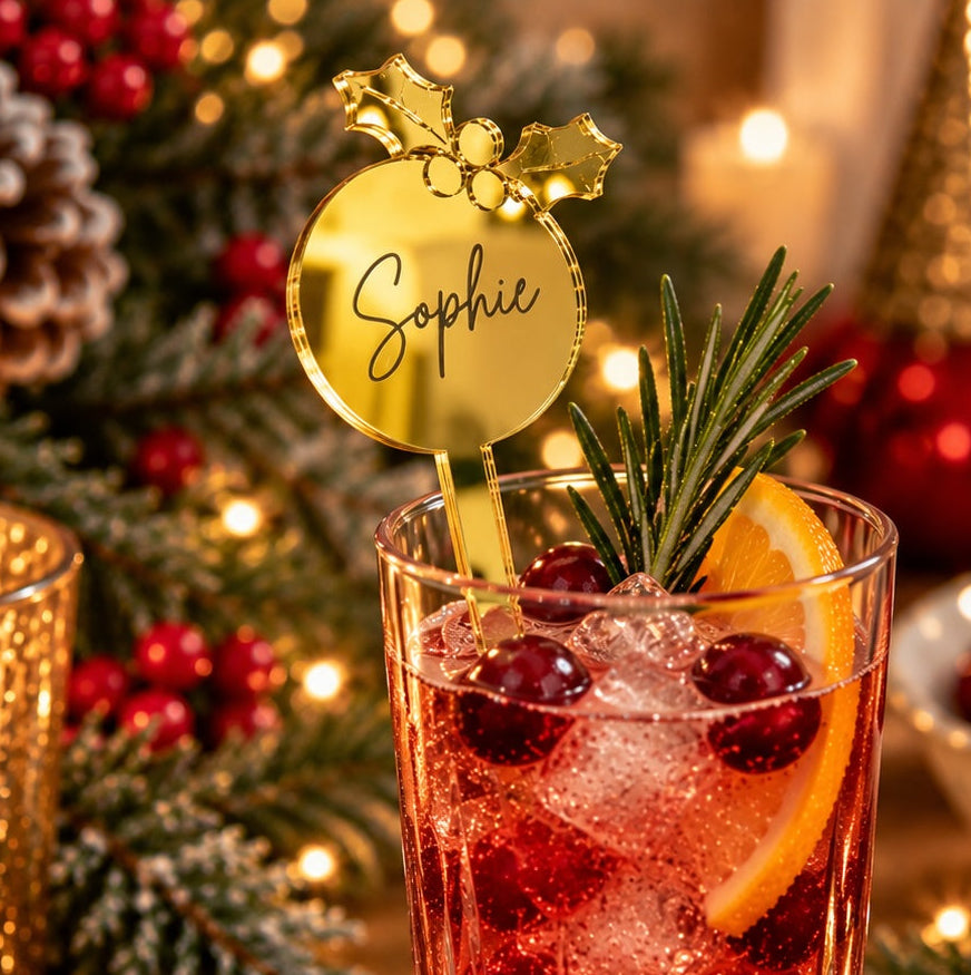 A festive holiday cocktail in a clear highball glass, filled with red drink, ice, cranberries, and an orange slice, with a gold sign reading 'Sophie' and a rosemary sprig garnish. The glass sits on a wooden table surrounded by Christmas decor, including pine branches, red ornaments, a lit candle, gingerbread cookies, and a tree in the background.