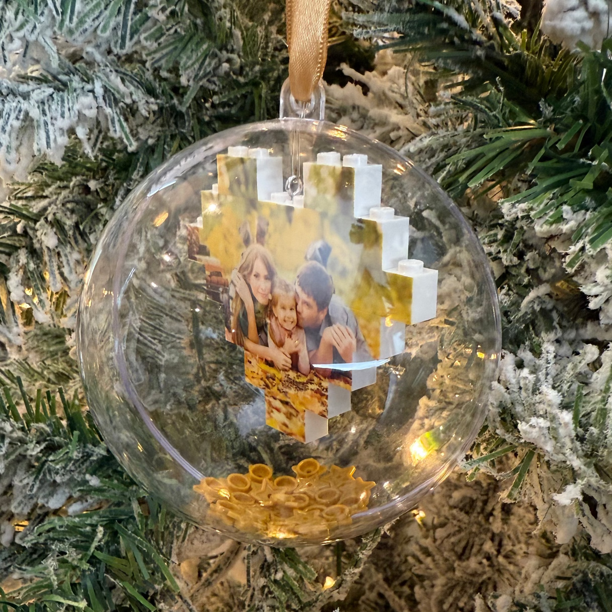 Clear Christmas ornament with a lego photo heart inside with gold stars, hanging on a decorated tree.