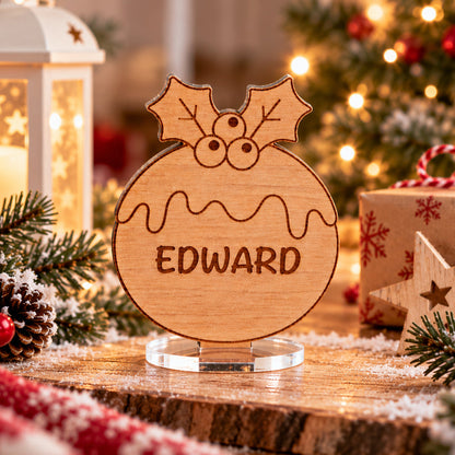 A personalized wooden Christmas ornament sign engraved with the name 'EDWARD' and holly leaves, displayed on a clear acrylic stand in a festive Christmas setting with lights, gifts, and candles.