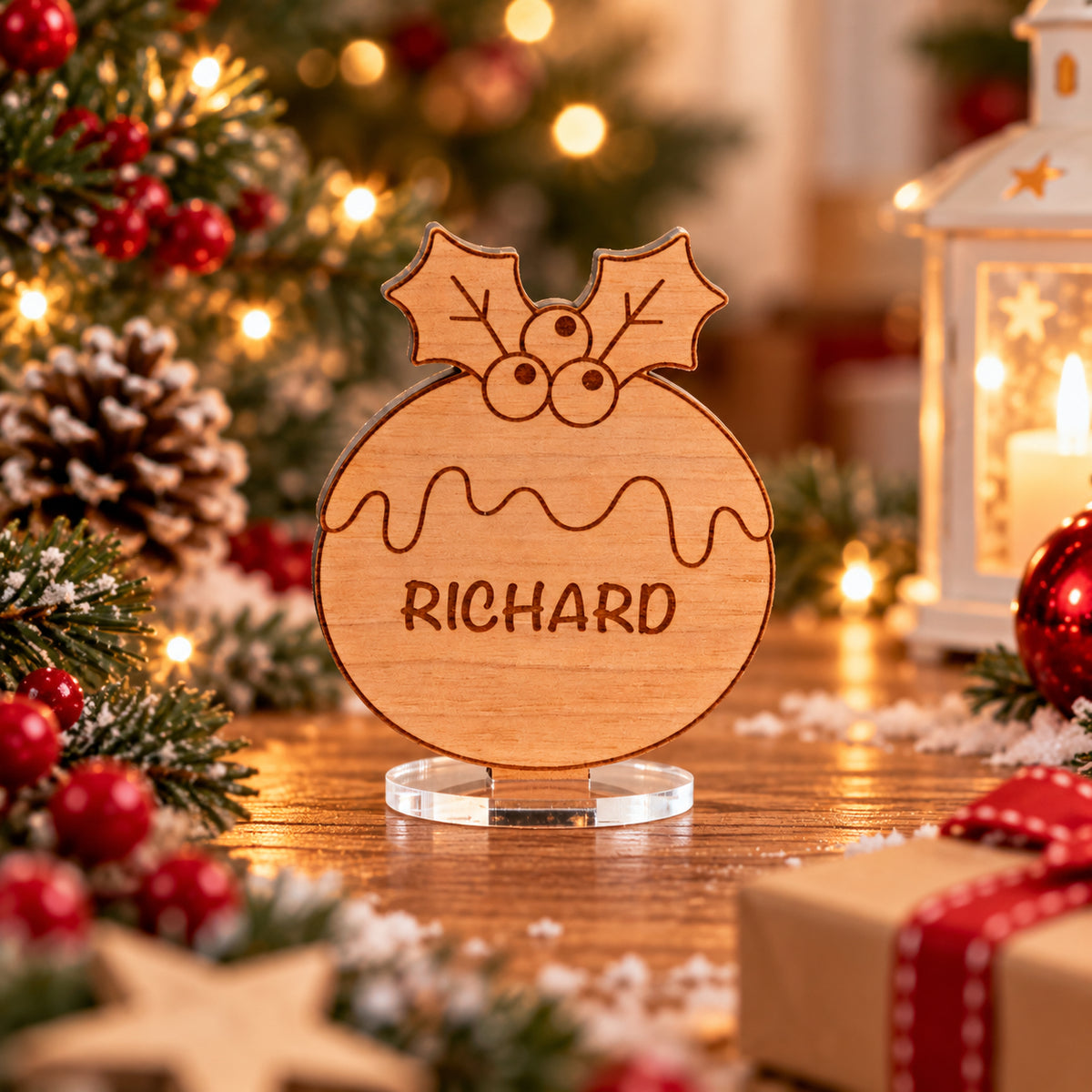 A personalized wooden Christmas pudding sign engraved with the name 'RICHARD' and holly leaves, displayed on a clear acrylic stand in a festive holiday setting with lights, pine branches, and ornaments.