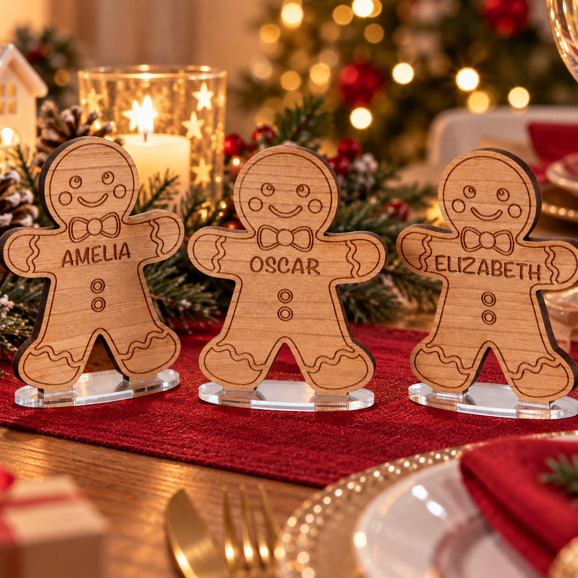 Three personalized gingerbread man table place cards on clear acrylic stands, engraved with the names Amelia, Oscar, and Elizabeth, set on a red table runner with Christmas decorations in the background.