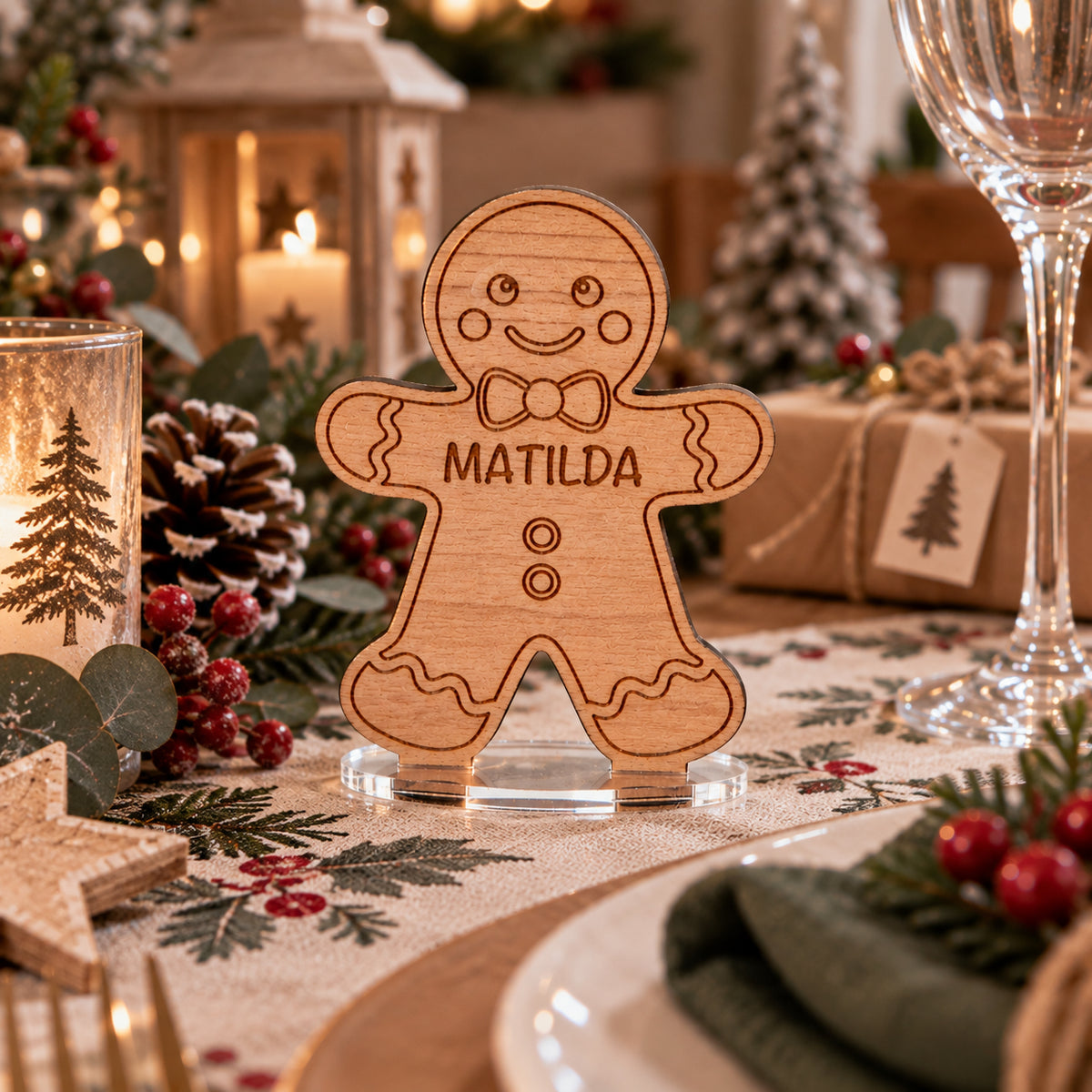 A personalized wooden gingerbread man table sign with the name 'MATILDA' engraved in the center, displayed on a decorated Christmas table runner with candles, pinecones, and wrapped gifts in the background.