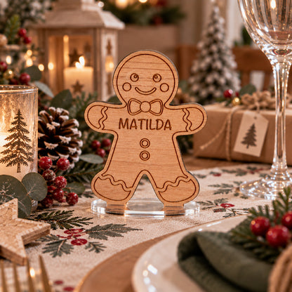 A personalized wooden gingerbread man table sign with the name 'MATILDA' engraved in the center, displayed on a decorated Christmas table runner with candles, pinecones, and wrapped gifts in the background.