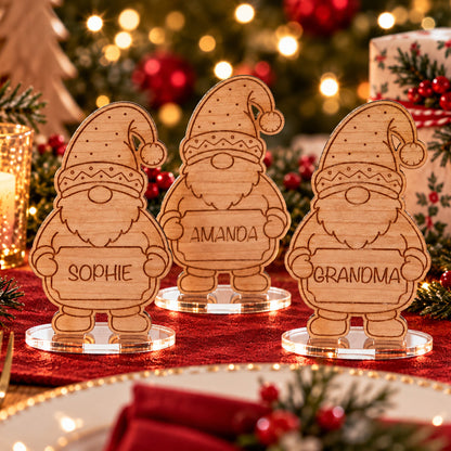 Three personalized wooden Christmas gnome place cards on clear acrylic stands, engraved with the names Sophie, Amanda, and Grandma, displayed on a red tablecloth with holiday lights, pine branches, and wrapped gifts in the background.