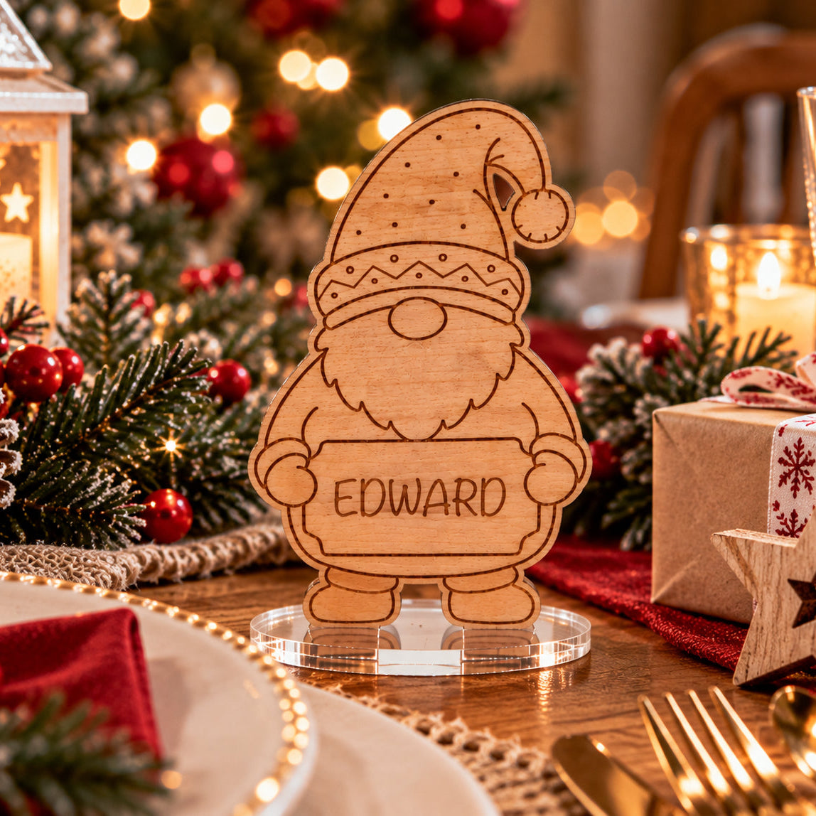A personalized wooden Christmas gnome stand engraved with the name 'EDWARD' on a clear acrylic base, set on a decorated Christmas table with pinecones, candles, and wrapped gifts.