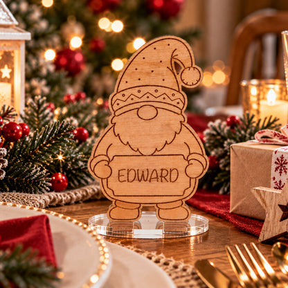 A personalized wooden Christmas gnome stand engraved with the name 'EDWARD' on a clear acrylic base, set on a decorated Christmas table with pinecones, candles, and wrapped gifts.