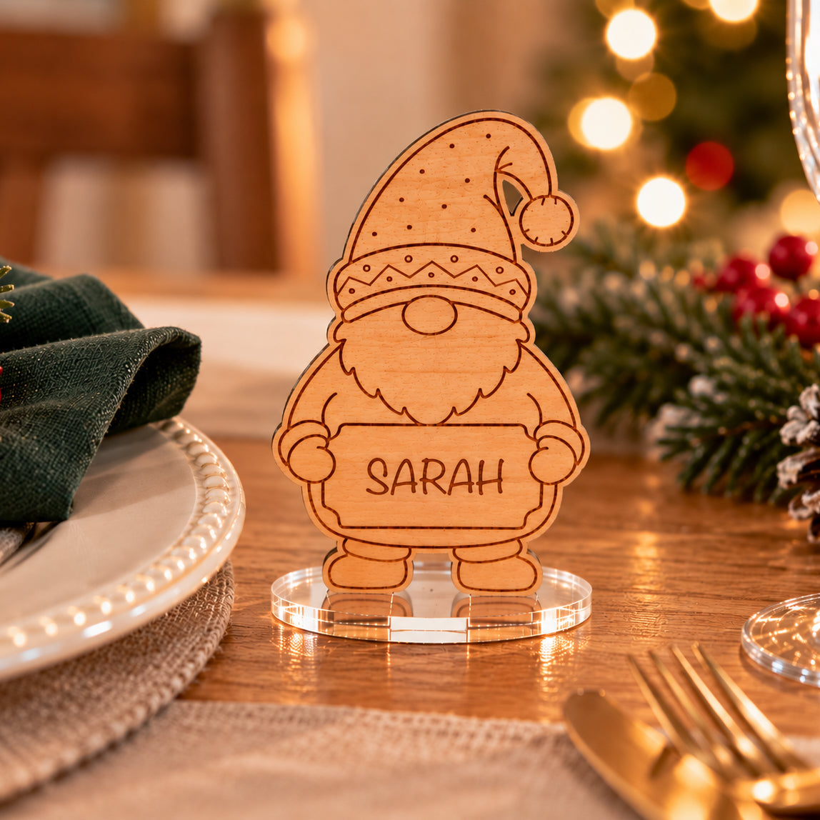A personalized wooden Santa place card stand on a clear acrylic base, engraved with the name 'SARAH', set on a decorated holiday table with a green napkin, gold cutlery, and a blurred Christmas tree in the background.