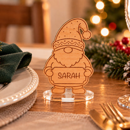 A personalized wooden Santa place card stand on a clear acrylic base, engraved with the name 'SARAH', set on a decorated holiday table with a green napkin, gold cutlery, and a blurred Christmas tree in the background.