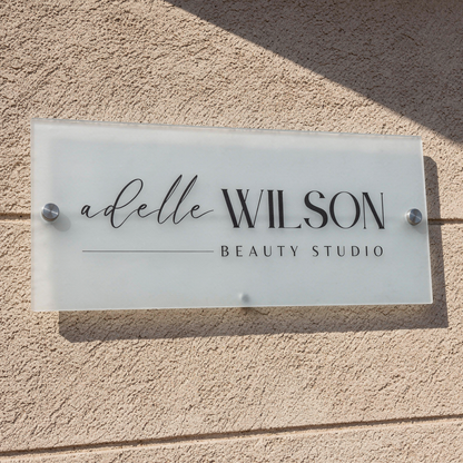 Frosted acrylic  sign with 'adelle WILSON BEAUTY STUDIO' on a textured wall.